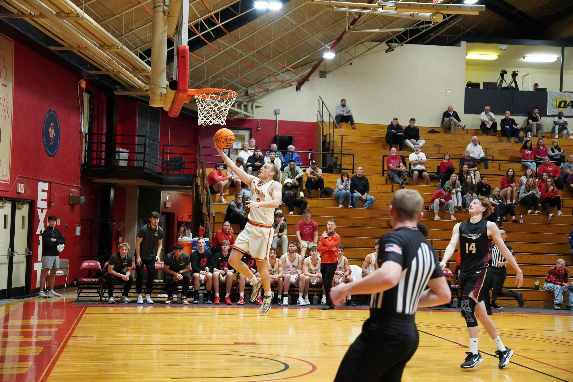 Jack Clement, an Otterbein basketball player goes for a layup as Jackson Rhodes, an Earlham College player looks on. A referee's back is towards the camera.