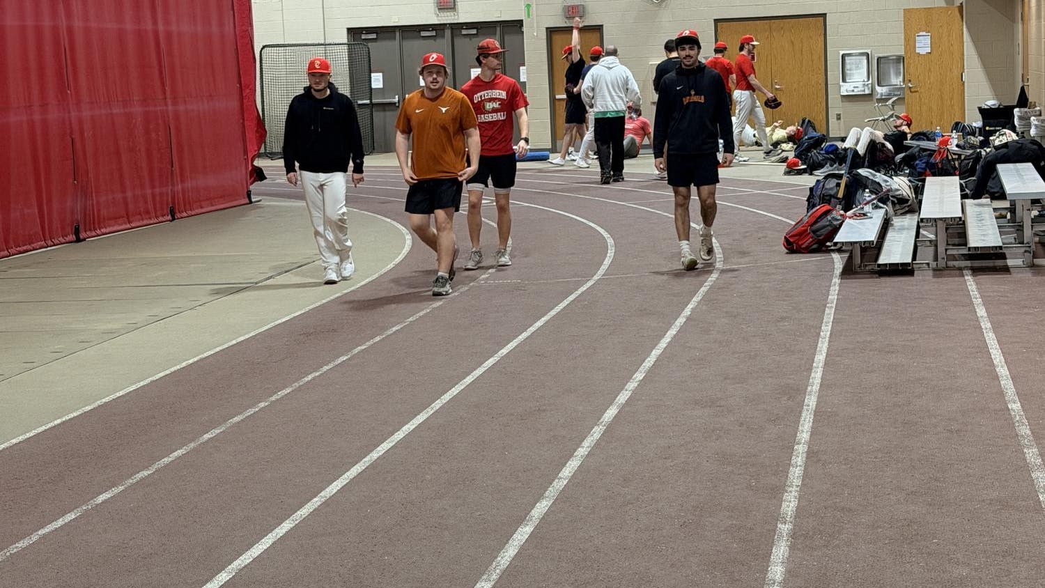 A group of four students in baseball caps walks towards the camera, and more students can be seen exercising in the background.