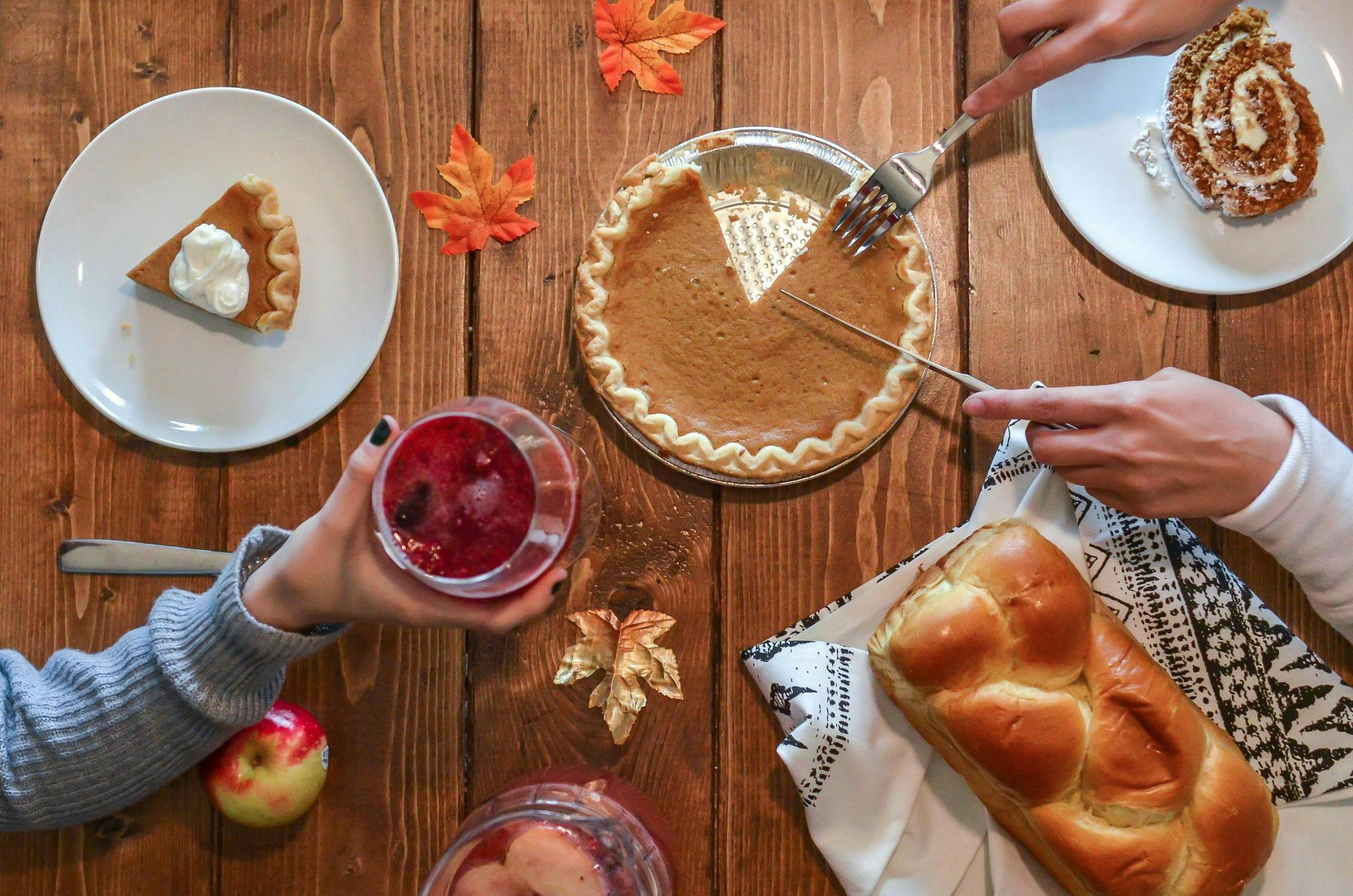 A wooden table with fall leaves and desserts. A hand is reaching over and holding a cup. Another pair of hands is slicing a pumpkin pie.
