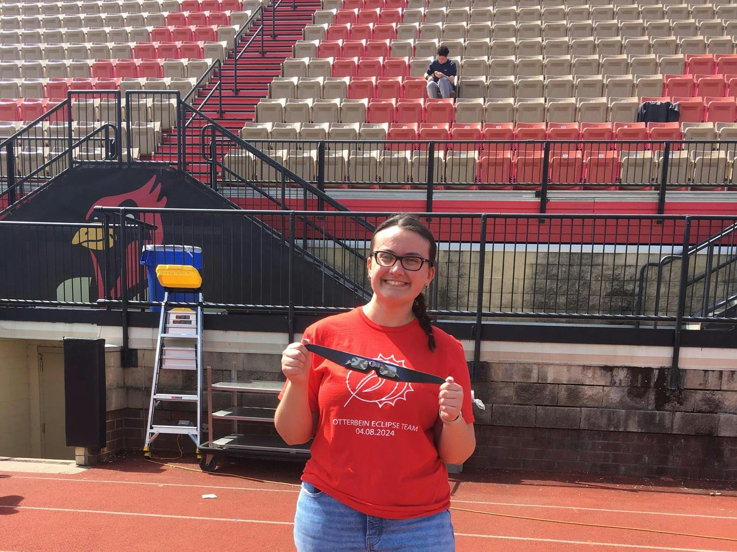 Girl in a red shirt that reads 'OTTERBEIN ECLIPSE TEAM 04.08.2024' in white lettering. She is smiling and holding up a pair of solar eclipse sunglasses.