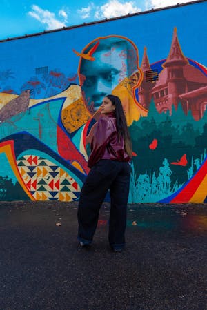Dark haired girl wearing jeans and a purple jacket posing away from the camera in front of an art wall.