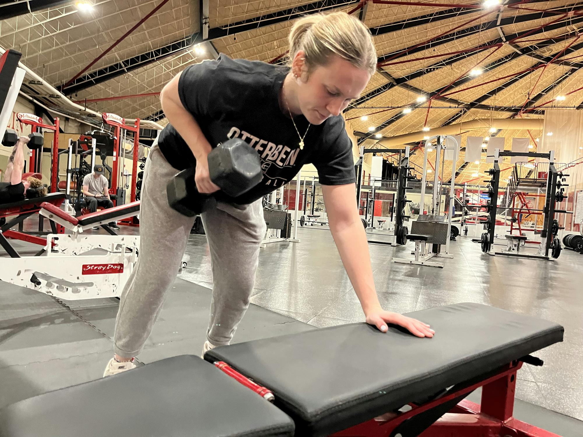 Blonde haired girl wearing a black shirt and grey sweatpants lifting a dumbbell. 