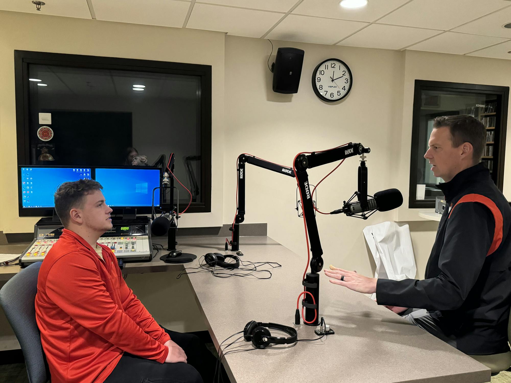 A reporter in red sits on the left side of a table while talking with Tom Snyder, who is dressed in black and sitting on the right. 