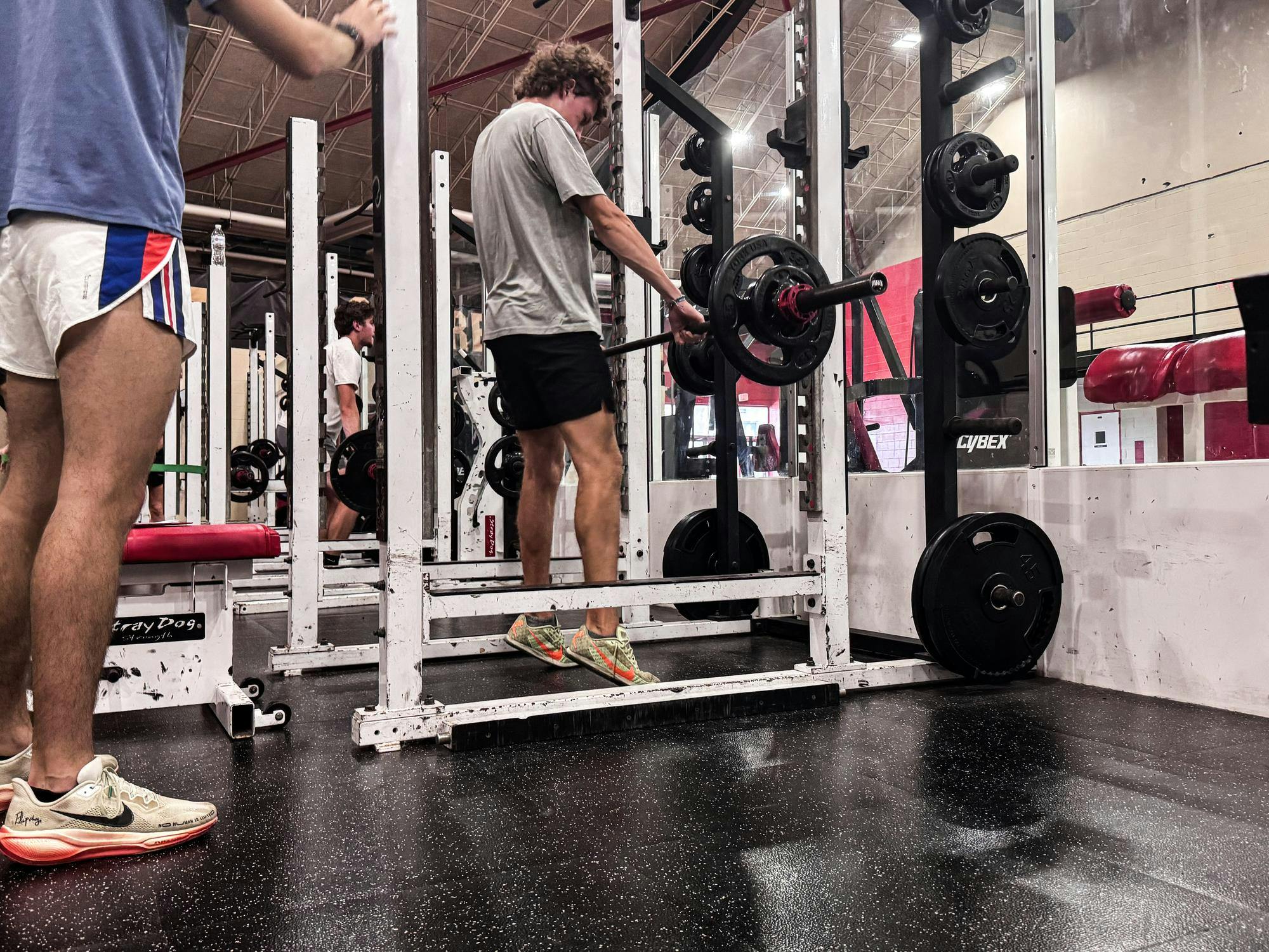 Man is lifting a barbell from the rack in the weight room with other teammates around him