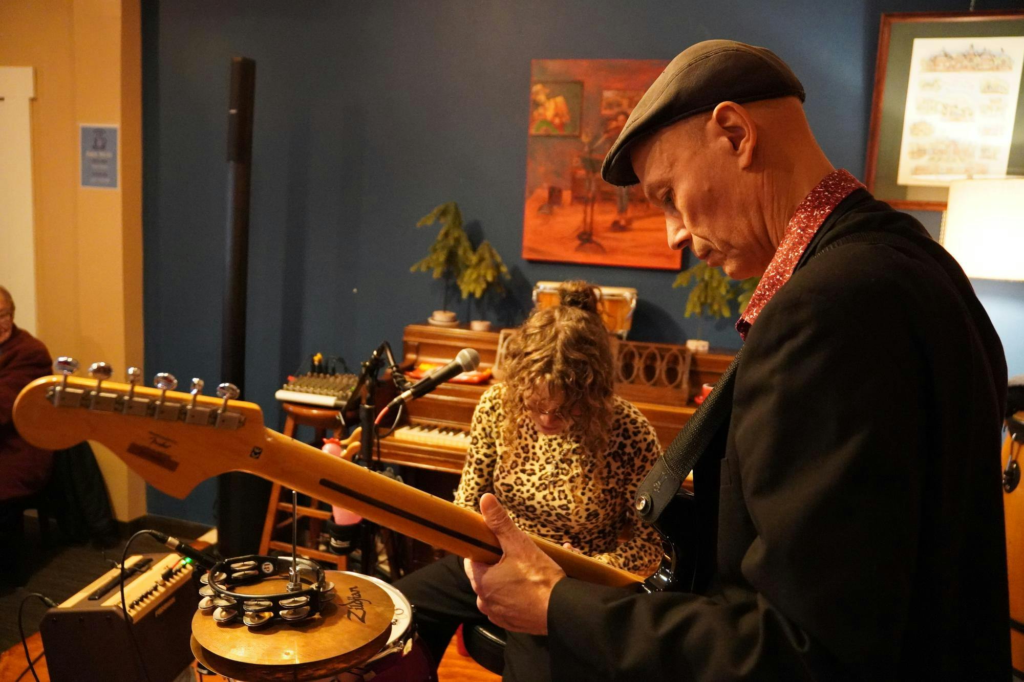 Woman sitting in front of a pink drum and symbol and a man tuning a guitar.