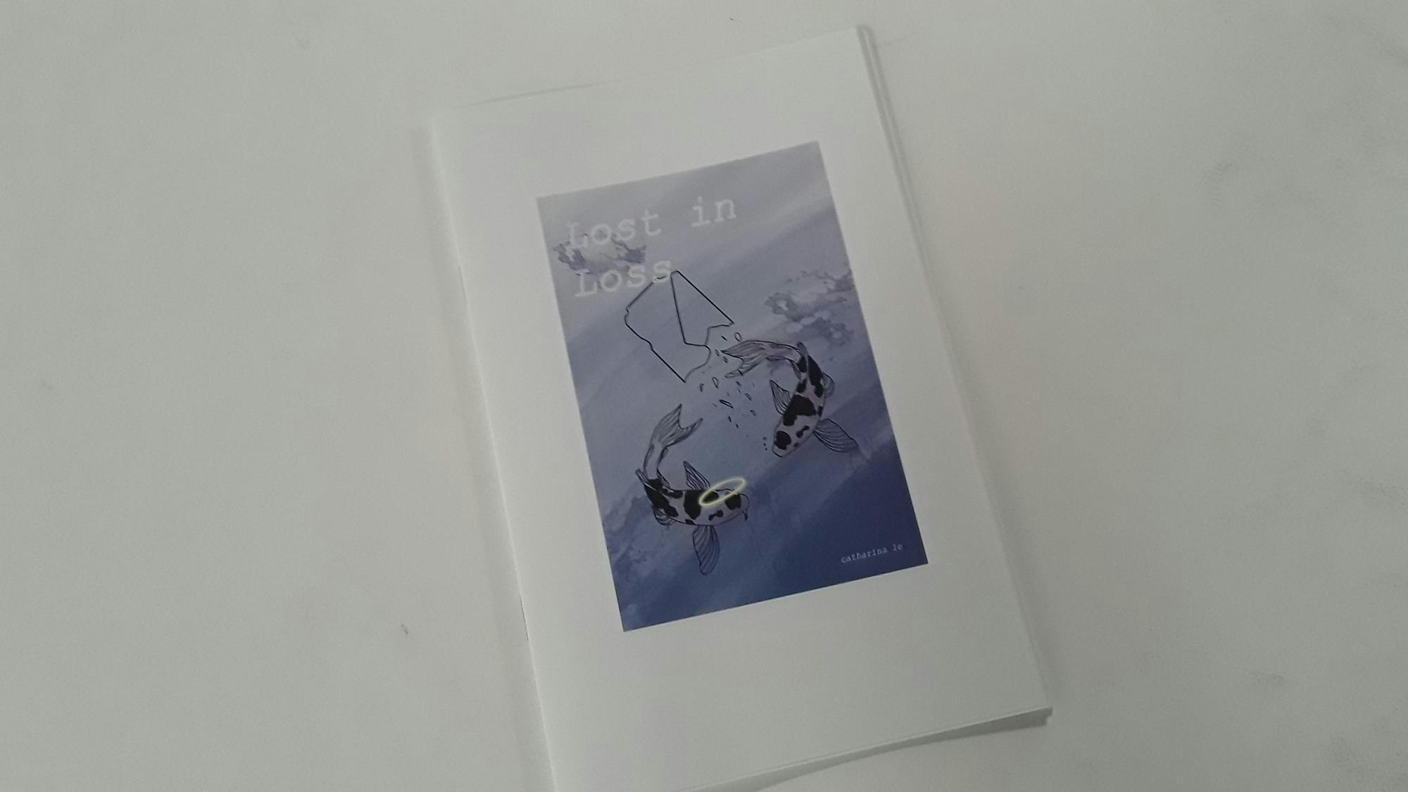 A thin stapled booklet. A centered image on the cover shows the title "Lost in Loss" and the author "Catharina Le." There are two koi fish swimming in water, one of which has a halo.