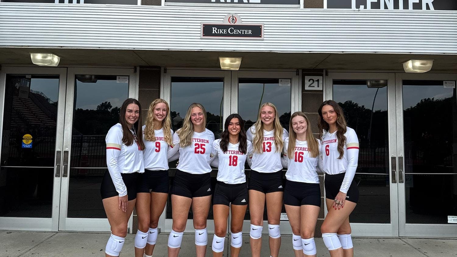 A group of women in volleyball jerseys pose in front of the Rike Center.