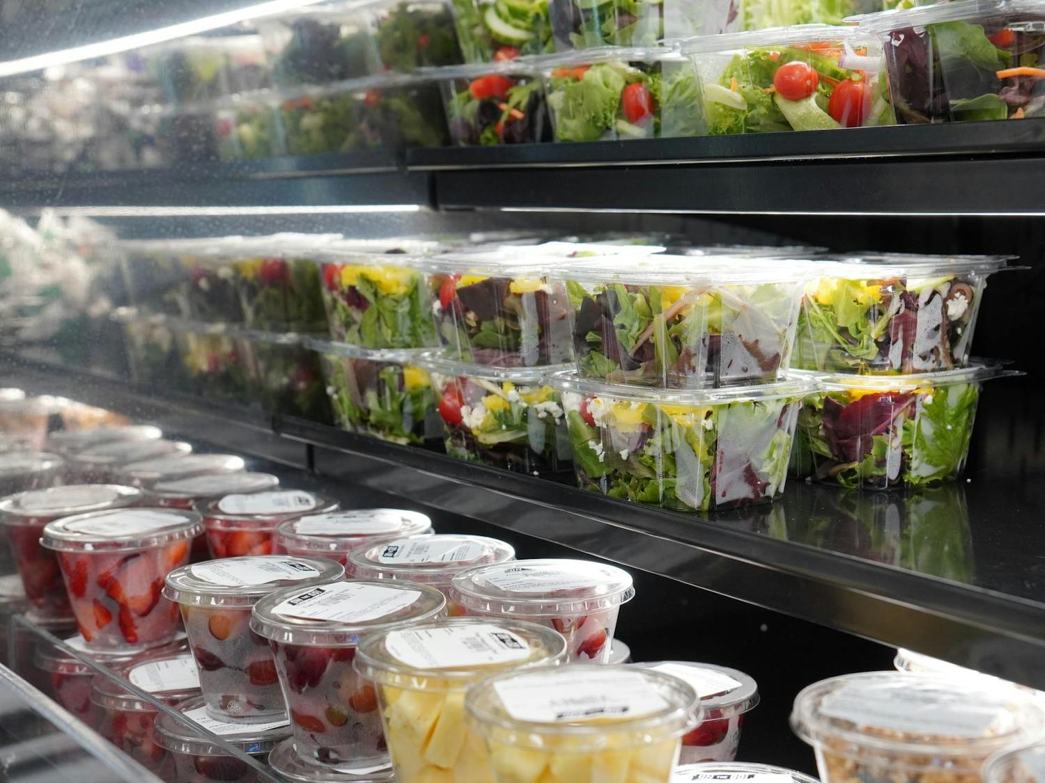 Pre-packaged containers of fruits and salads are stacked and placed in rows on shelves inside a cooler. 