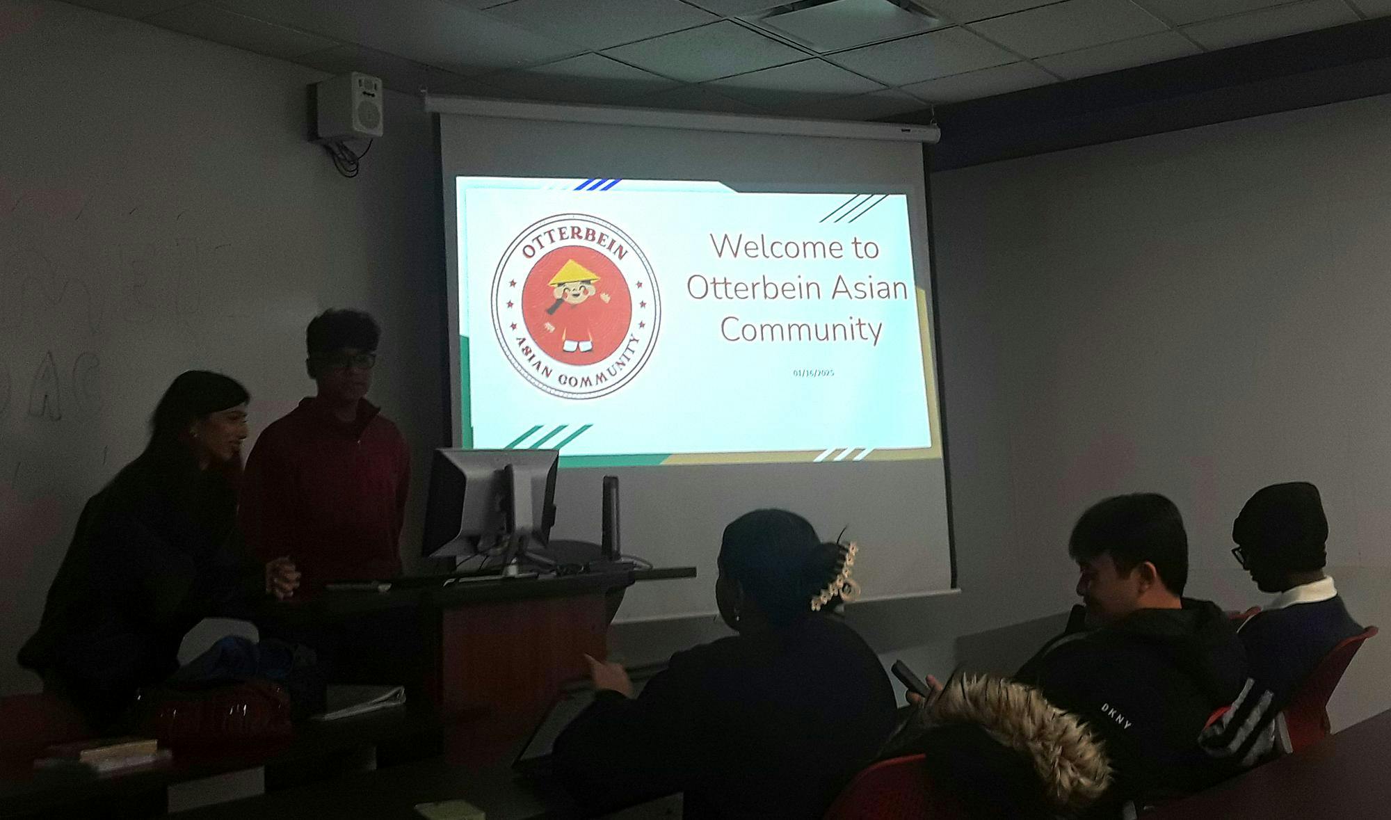 Two students stand behind a podium in front of a row of students seated at a table in the front row of a classroom. Projected on the screen is a slideshow that says "Welcome to Otterbein Asian Community."