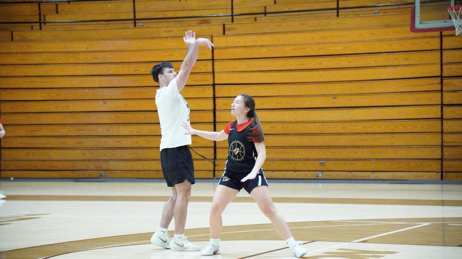 The photo depicts two individuals on a basketball court. One is a man in a white tee shirt and black shorts, while the other is a woman in a red shirt and a jersey over top with black shorts. Kadence Conley is putting out her arm in order to defend the shot from practice player Jordan Ermalovich. The players are slightly smiling.