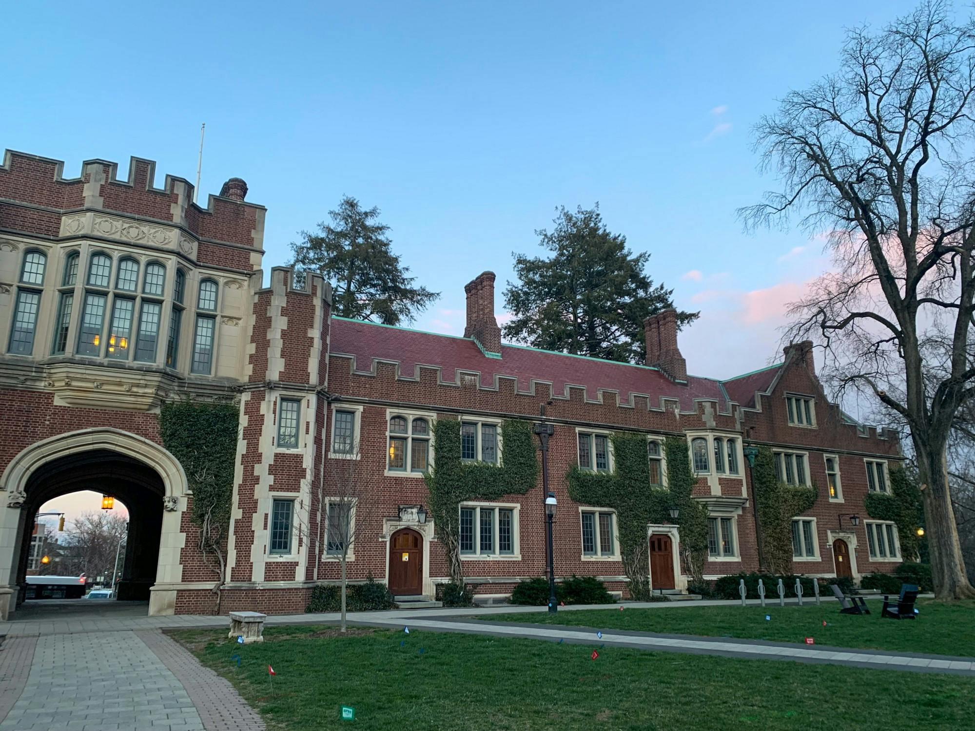 Red brick building with ivy growing between some of its windows. The left side of the building has an arch with large bay windows above the arch. The sky is blue with wisps of pink clouds.