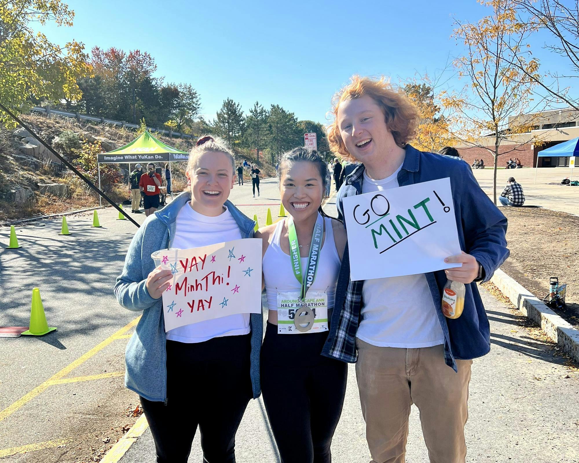 A woman in a white tank top with her hair pulled back smiles with people on either side of her, holding signs that say 'YAY MinhThi! YAY' and 'GO MINT.' A medal hangs from her neck by a green ribbon.