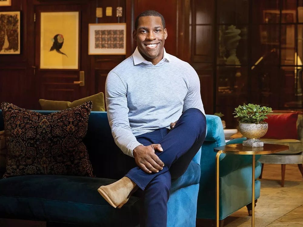 A Black man sitting on a couch with his legs crossed dressed in business casual attire. 