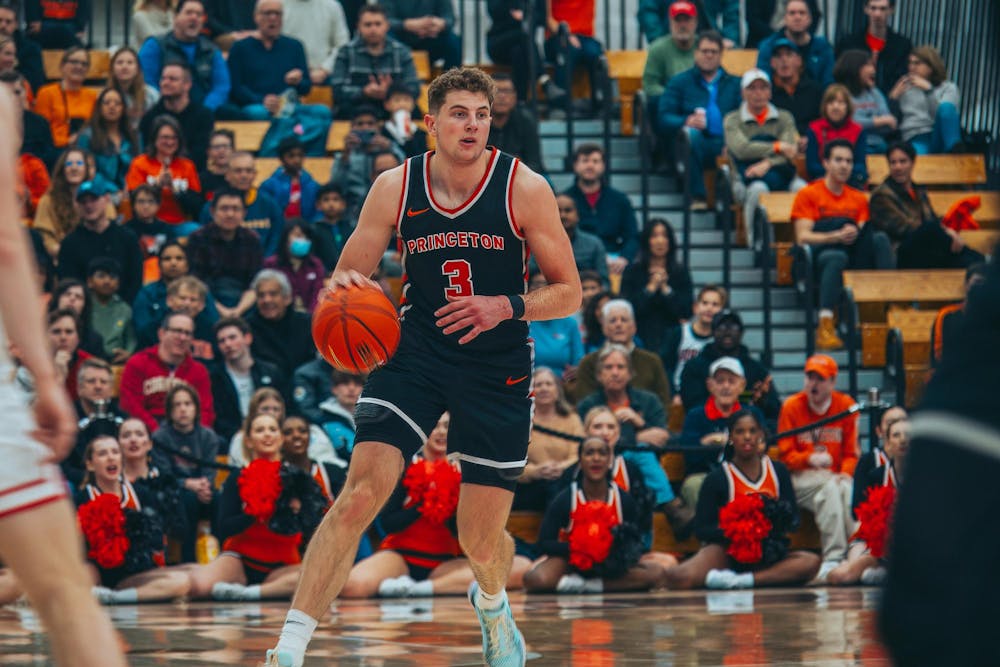 A man wearing black shorts and a black jersey with the number three, Princeton, and Nike logo on the jersey in orange dribbling a basketball on a court. 