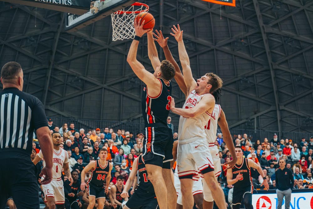 A basketball player wearing a black and orange jersey is mid-air, dunking a basketball into a hoop. Two defending players wearing white and red jerseys attempt to block the shot.
