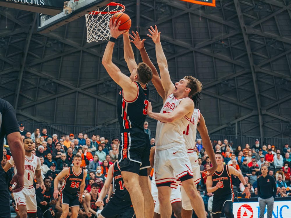 A basketball player wearing a black and orange jersey is mid-air, dunking a basketball into a hoop. Two defending players wearing white and red jerseys attempt to block the shot.