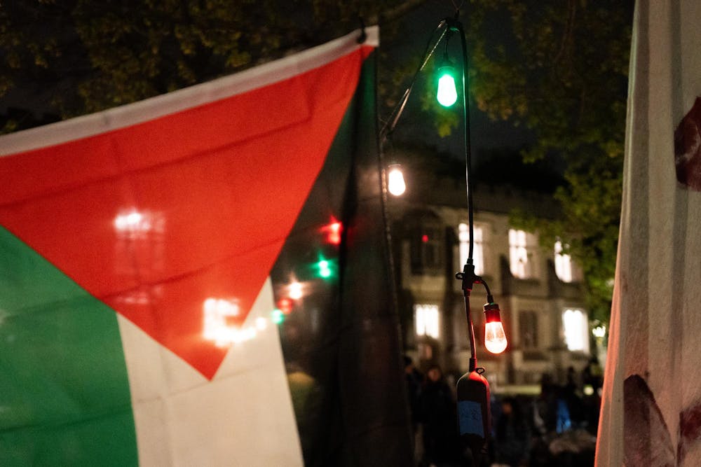 Red, white, green, and black flag in the foreground. In the background, multicolored lights and a stone building.