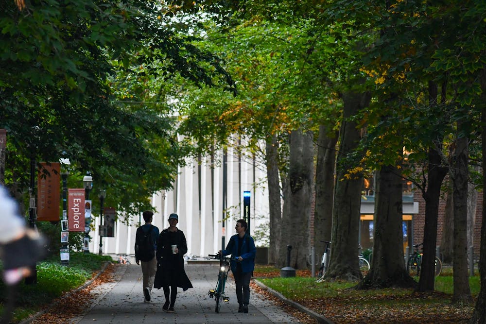 Three students with backpacks walk down a tree-lined path on campus in front of the SPIA building. 