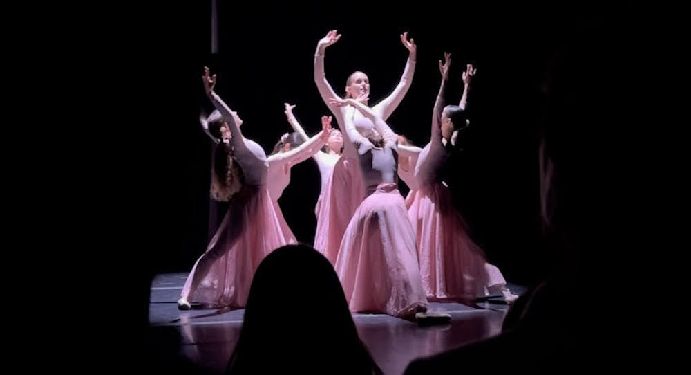 Seven dancers in long, flowy pink dresses with their hands outreached upwards.