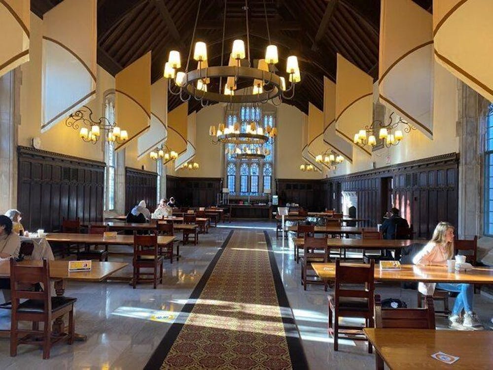 An image of a dining hall with high ceilings adorned with chandeliers. Wooden tables line each side of the room. Sunlight streams into the building. Students are seated one to a table and social distancing signings are present on the floor. 