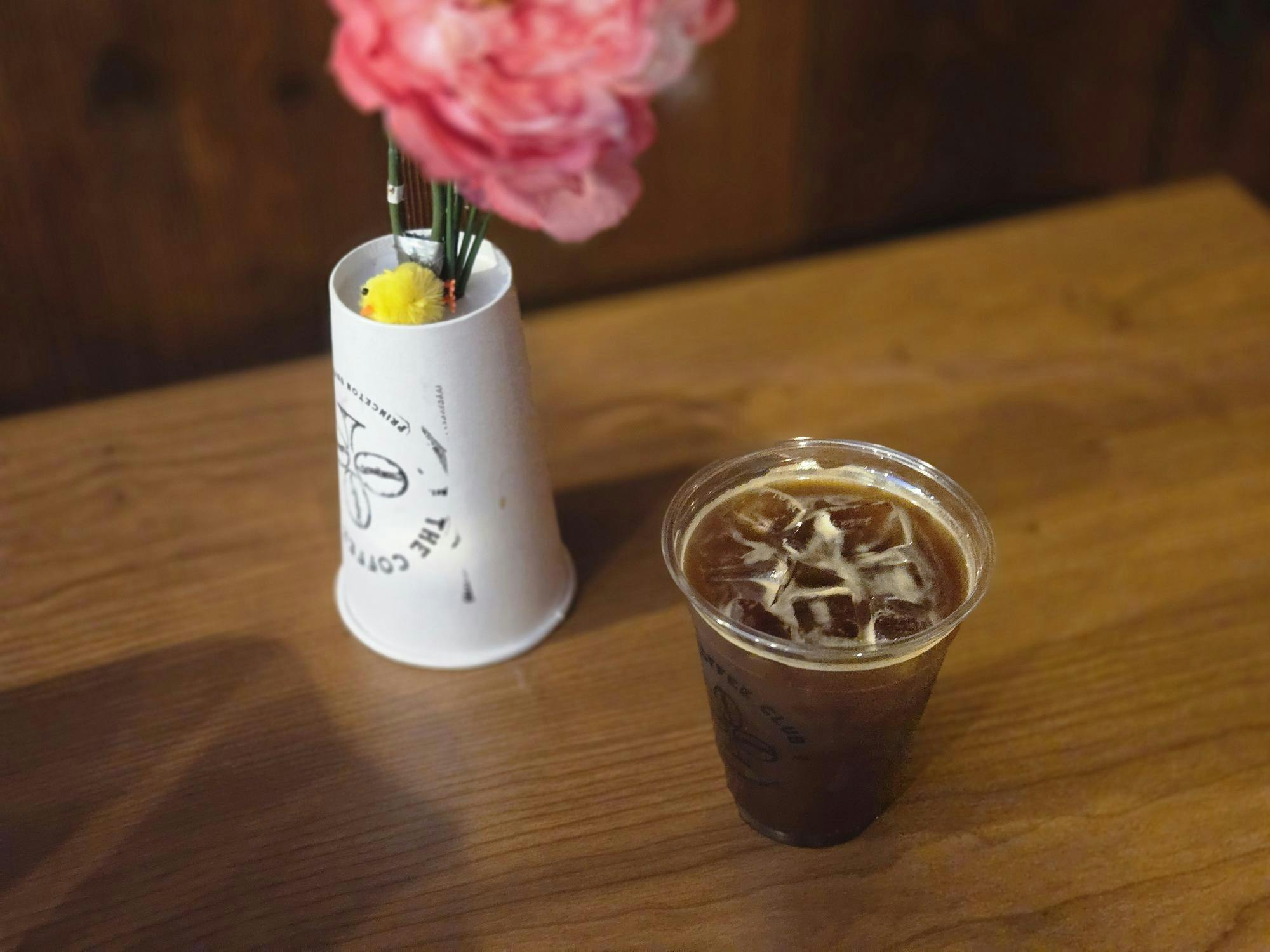 A brown iced drink in a transparent cup and a white Coffee Club cup that is upside down sit on a brown table.