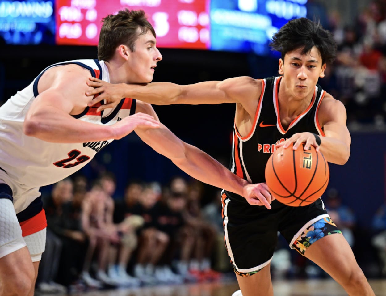 Princeton player wears a black jersey as he dribbles a basketball with his left hand. He is defended by a Duquesne player wearing a white jersey.