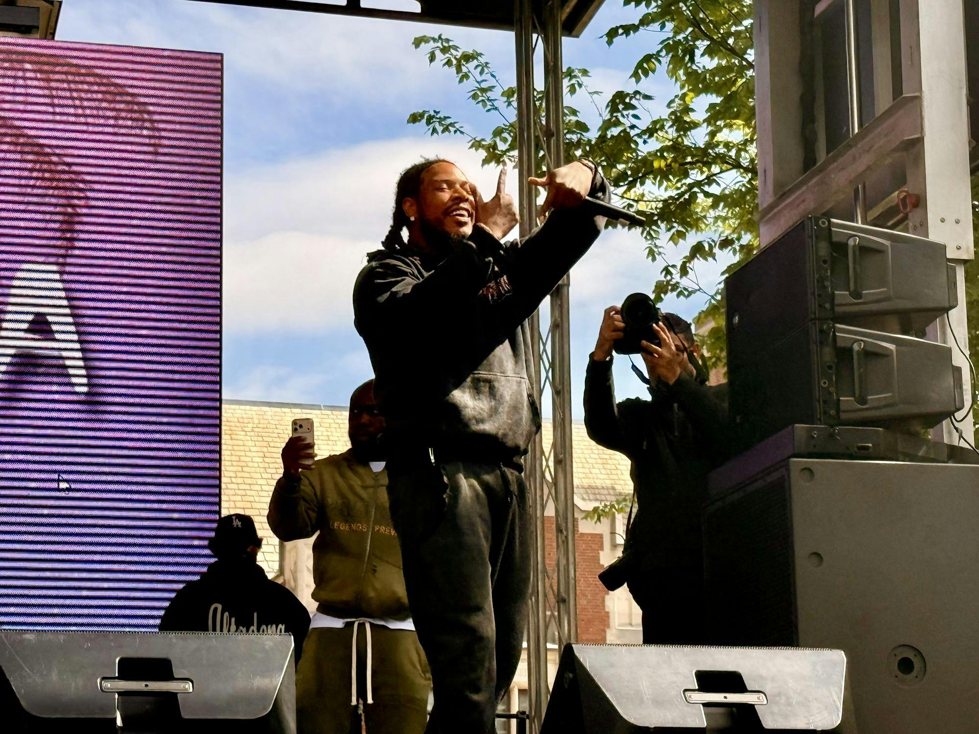 Man in black hoodie and black pants sings to a crowd behind a neon screen on a stage. 