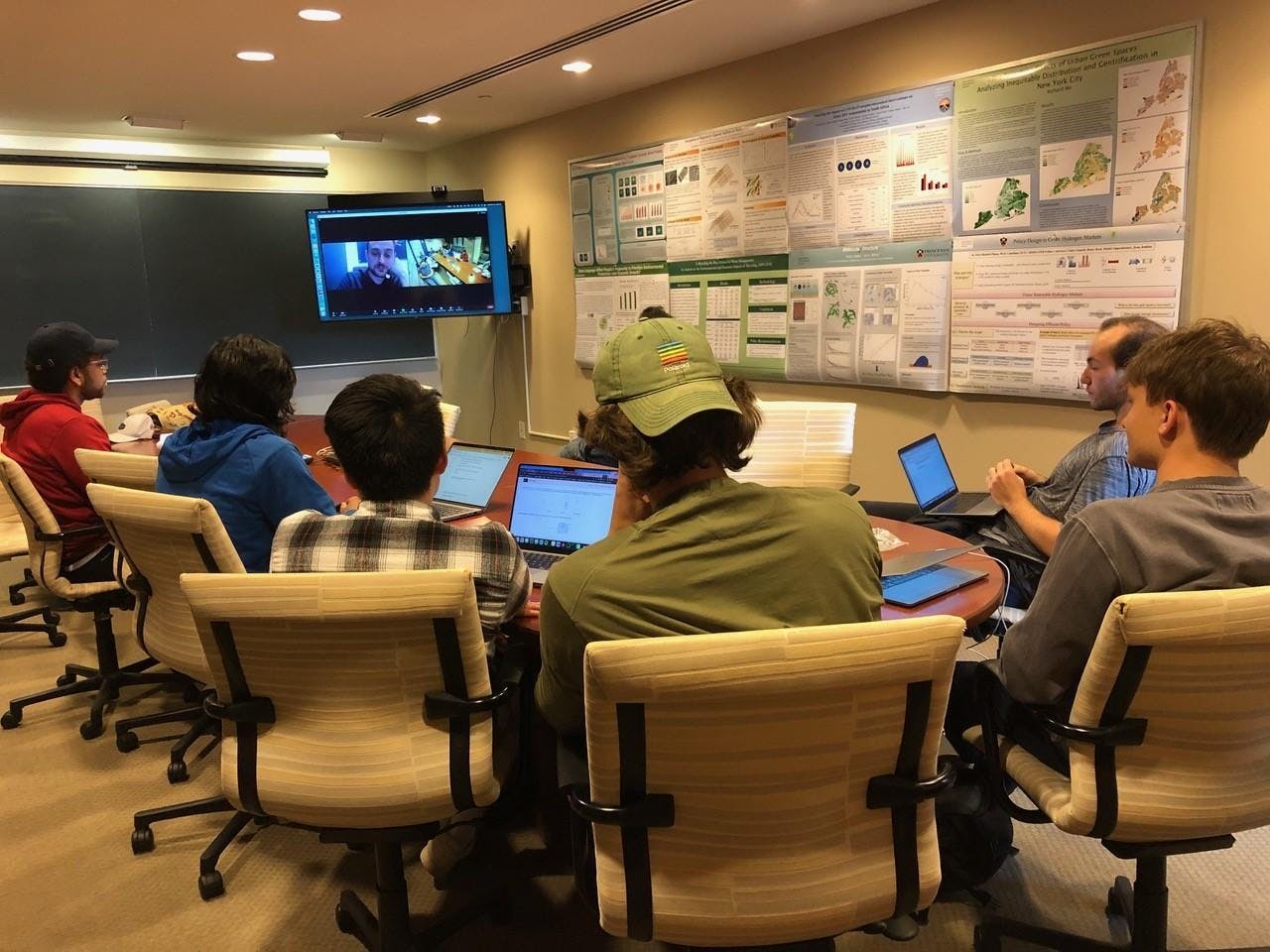 A group of people sitting around a table with computers in front of them inside a small room with a television inside. 