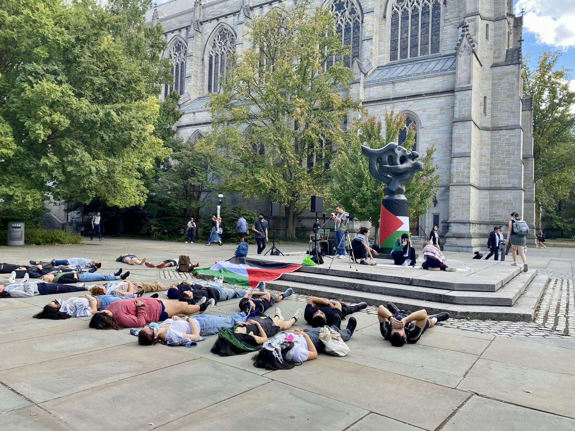 About 20 people lie flat on concrete in front of a dark gray statue wrapped in a black, red, white and green flag