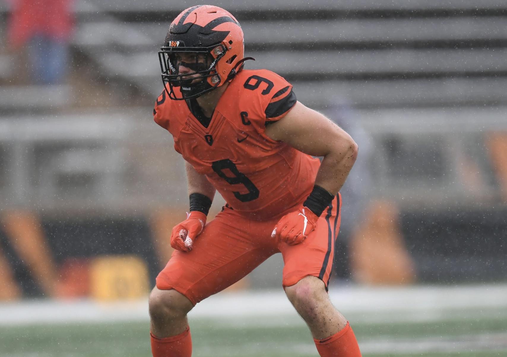 A football played in an orange and black uniform crouches on a field.