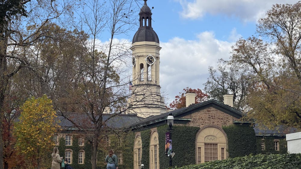 Nassau Hall in autumn, viewed from a downhill path with colorful trees framing its central clock tower; historic stone facade partially obscured by foliage, with two people facing away from the camera walking towards the building.