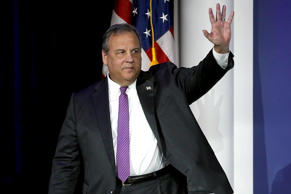 A man in a suit standing in front of an American flag waves. 