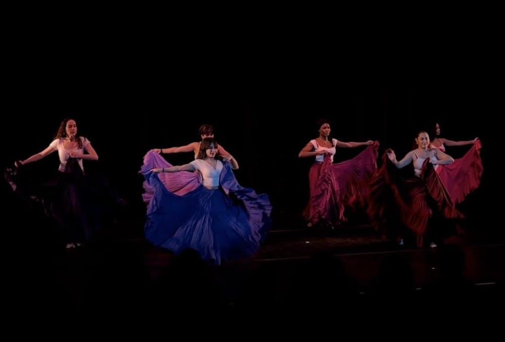 Five dancers in long purple and magenta skirts do a Cumbia and Mapalé style dance on stage.