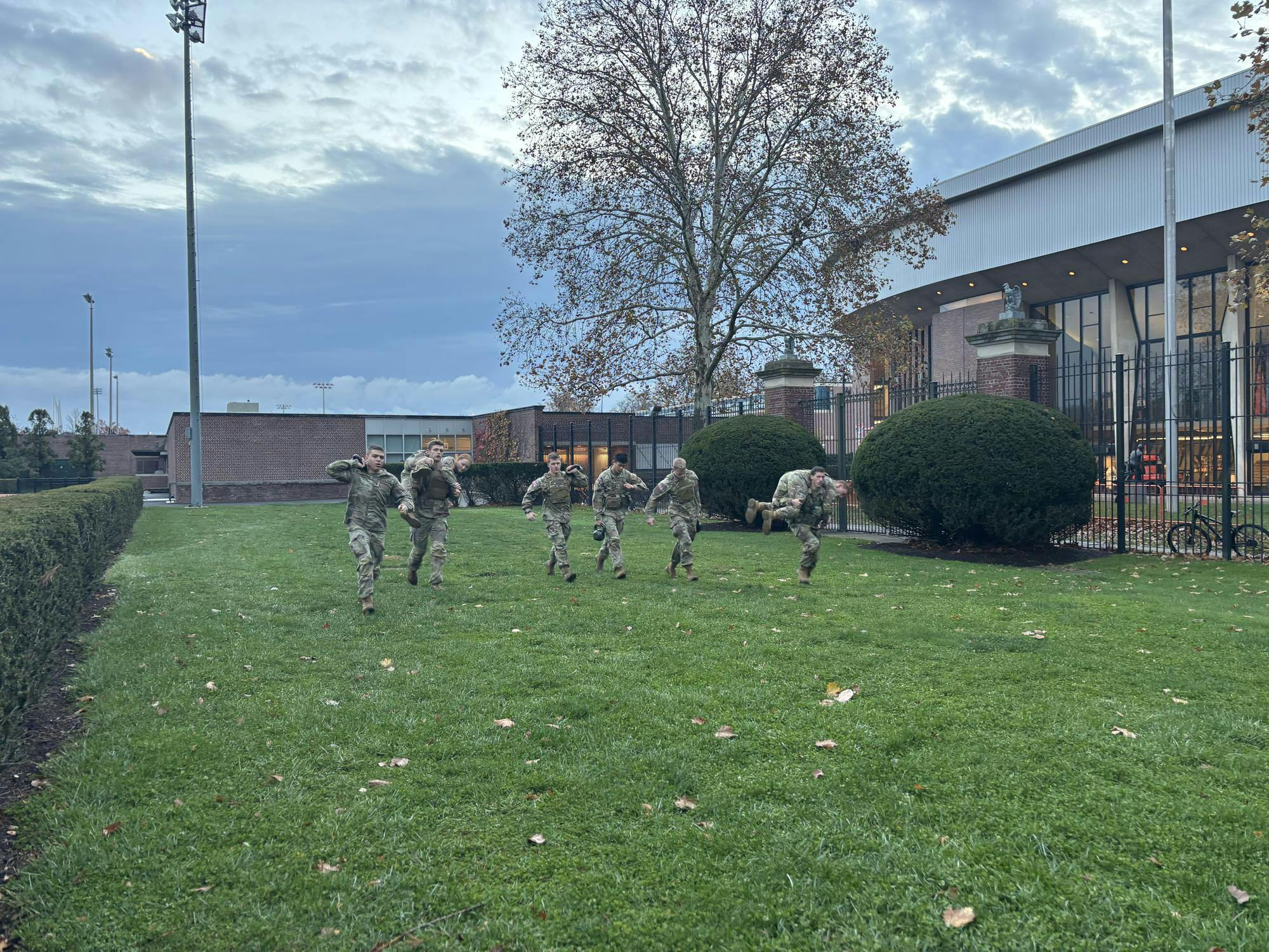 A group of ROTC cadets dressed in tan and olive green uniforms run across a grass field with a building next to them.