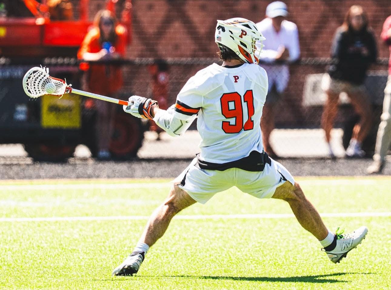 A man wearing white shorts and a white jersey holding a lacrosse stick in his left hand with a ball in it, preparing to shoot towards the goal.