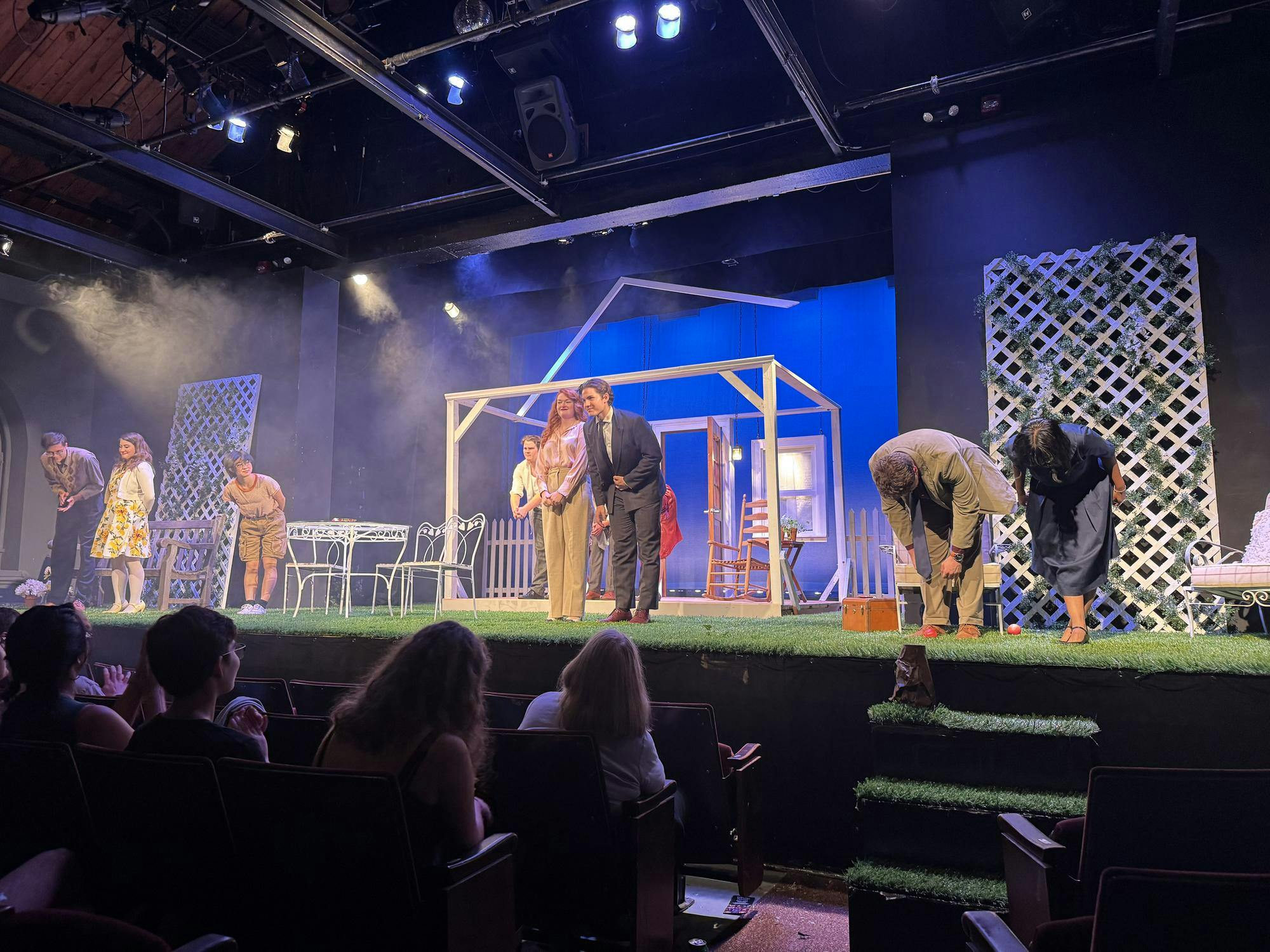 People in a multicolored outfits bow together. There is a green fake-grass covering over the stage.