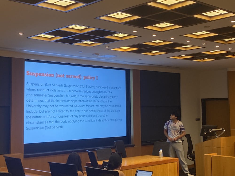 A young man wearing a white t-shirt stands in front of a smartboard, which has a presentation with the title “Suspension (not served): Policy I.”