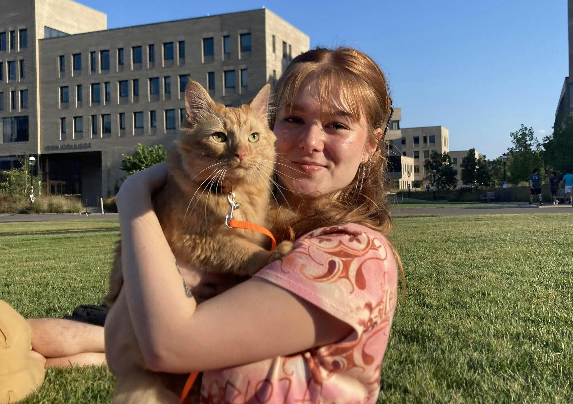 A young woman wearing a pink shirt holds a ginger cat in her arms on a sunny day. They sit on a grassy field with a rectangular light brick building in the background. 