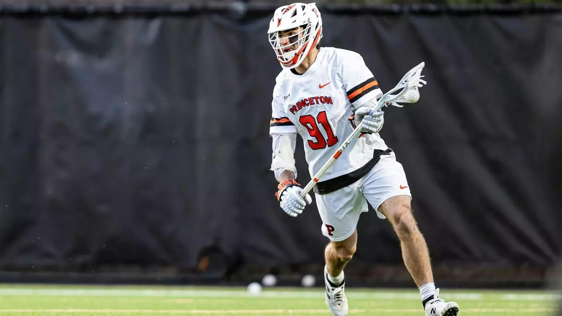 A man with white shorts and a white jersey holding a lacrosse stick with a ball in it on a grass field. 