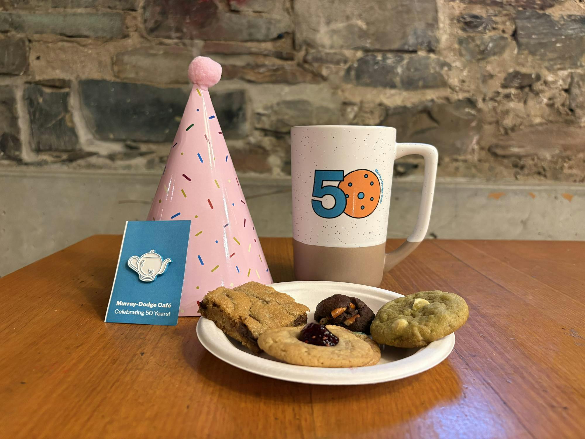 A mug, teapot pin, cookies, and a party hat rest on a wooden table.