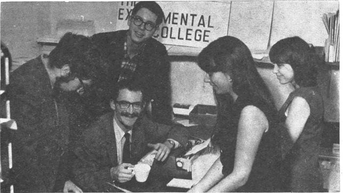 A black and white photograph of four students surrounding Daniel Altman ’67 seated at a desk holding a cup of coffee at the Experimental College.