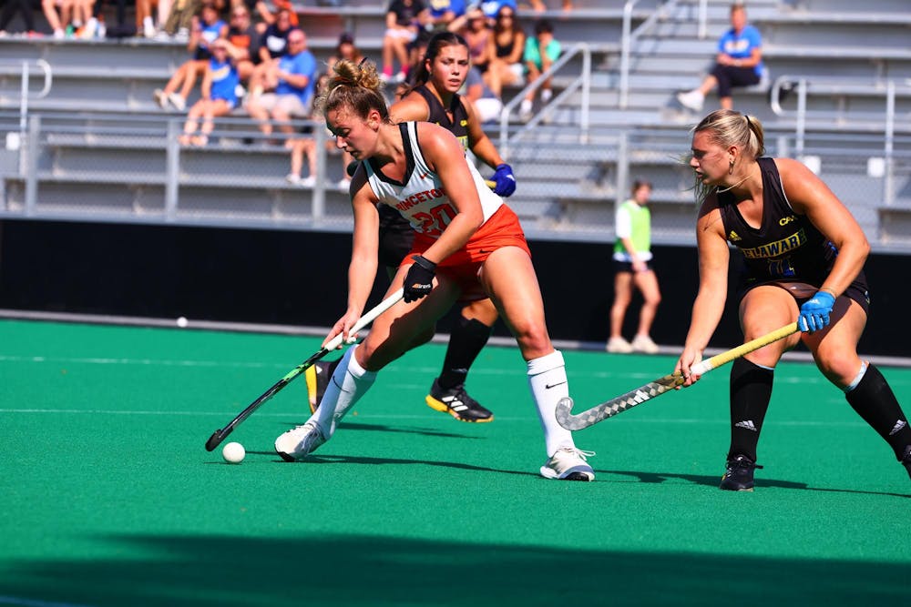 Woman in white field hockey uniform shoots while being guarded by woman in black uniform.