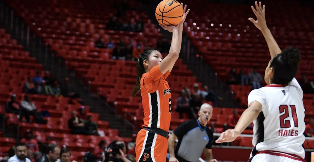 Girl in orange Princeton uniform shooting basketball over opponent in white, referee in back looking at the game with whistle in mouth. 