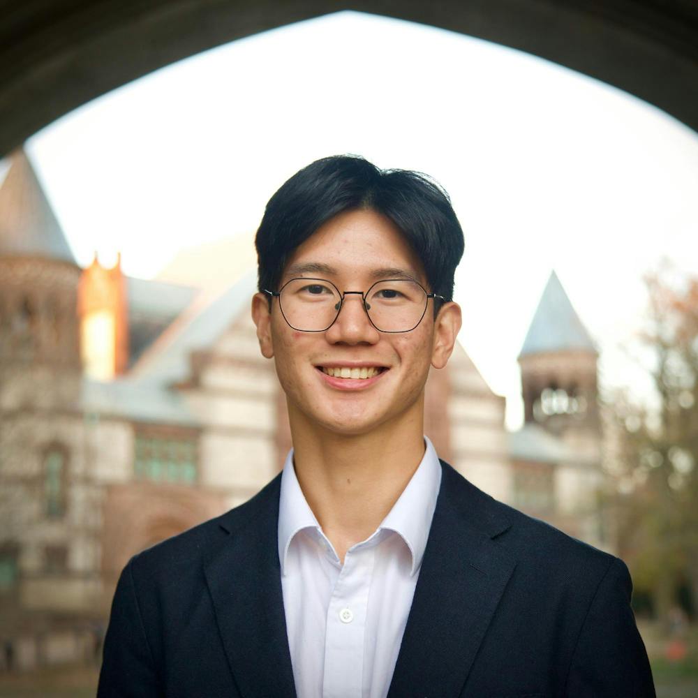 A man wearing a suit and glasses in the foreground, and a blurred brown building lays in the background.