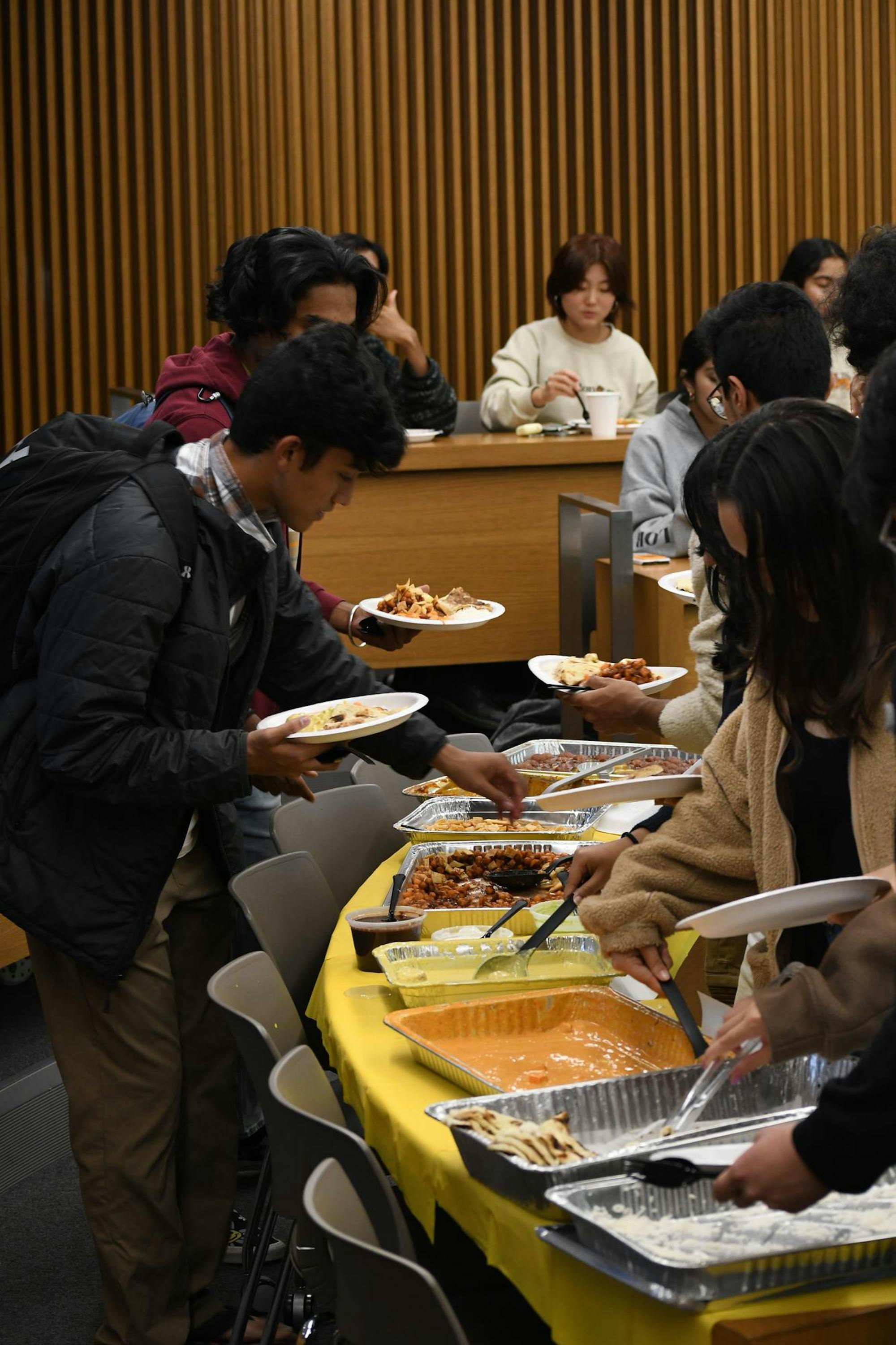 A number of students holding white plates of food gather around a table with silver food containers