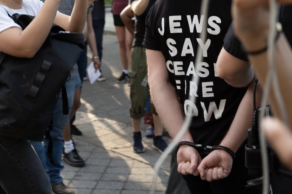 A man with a black shirt with white text that says "Jews say Ceasefire now" is in handcuffs.