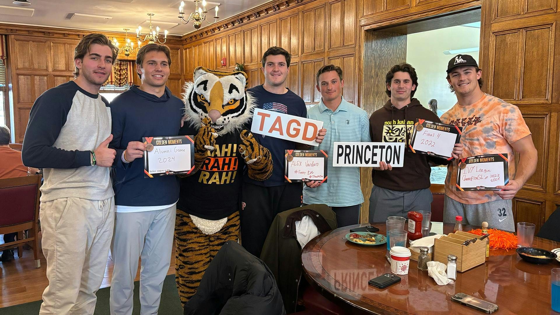 A group of men posing with a Tiger mascot in a dining room holding up signs. 
