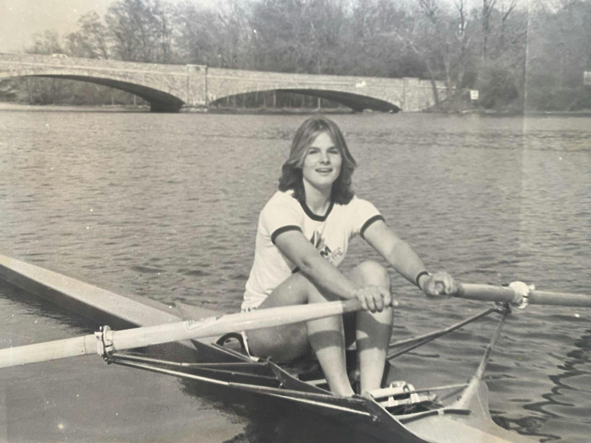 A woman sits in a boat in Lake Carnegie. There is water around her and a bridge in the background. The photo is black and white.