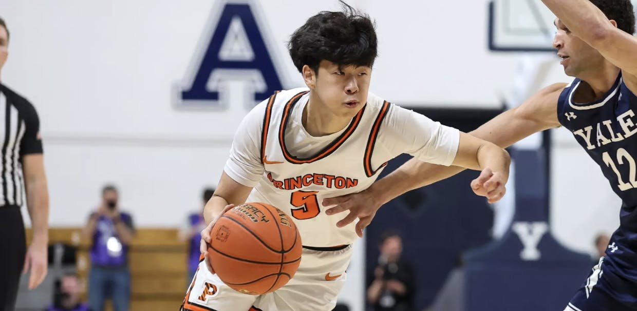 A Princeton player in a white jersey dribbling a basketball, being guarded by a player in a blue jersey.