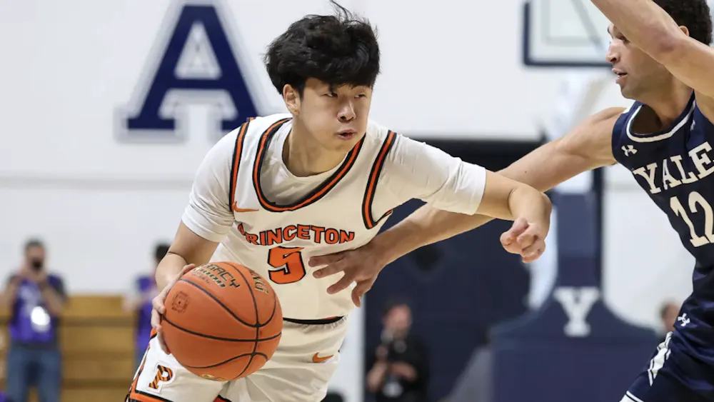 A Princeton player in a white jersey dribbling a basketball, being guarded by a player in a blue jersey.